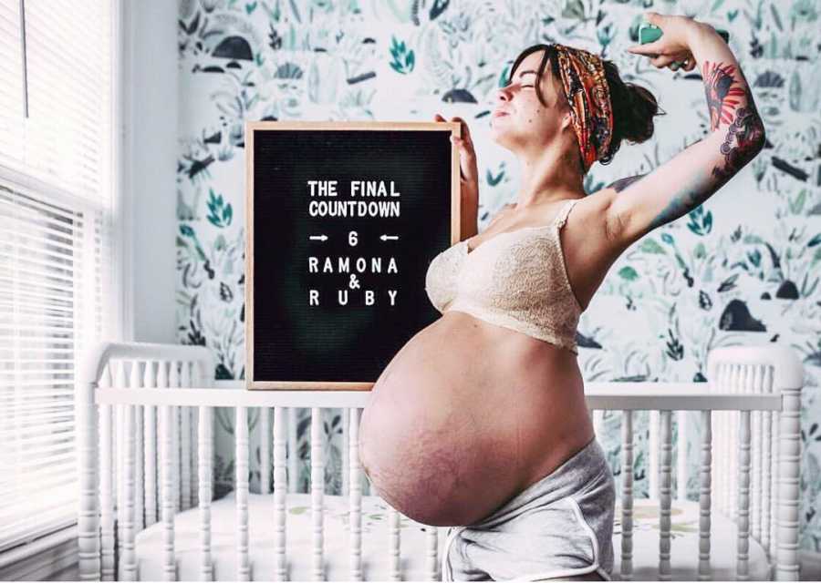 Woman pregnant with twins stands beside crib with sign that says, "the final countdown Ramona and Ruby"