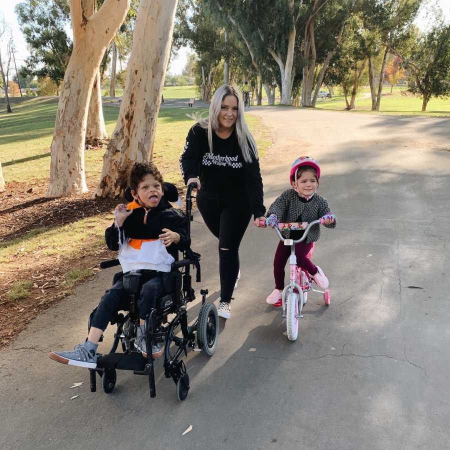 Mother smiles as she walks on path pushing son with Williams Syndrome in wheel chair and holds handle of daughter's bike