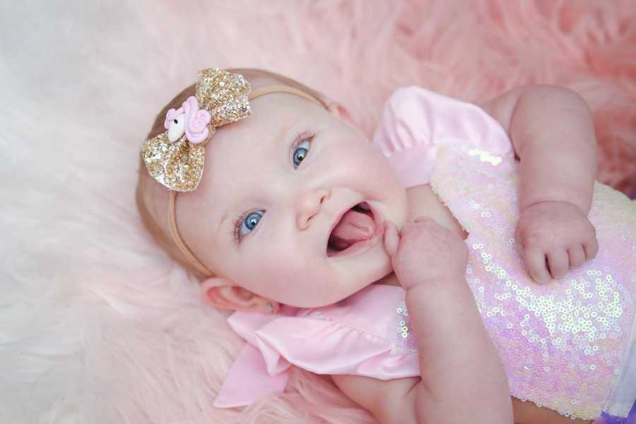 Baby girl lays on back in pink sparkly outfit while sticking tongue out