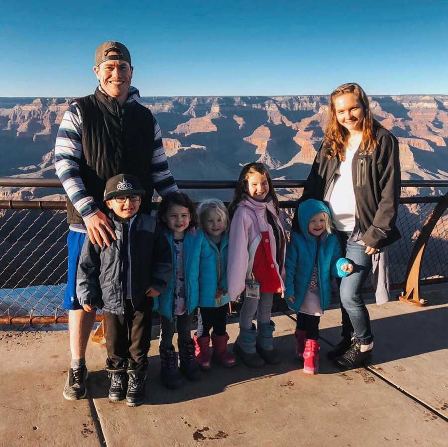 Husband and wife stand in front of fence that looks out to the Grand Canyon with their five young kids