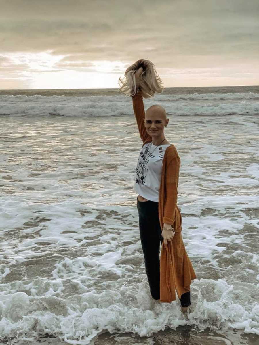 Woman with hodgkin's lymphoma stands on shore of beach holding her blonde wig in air