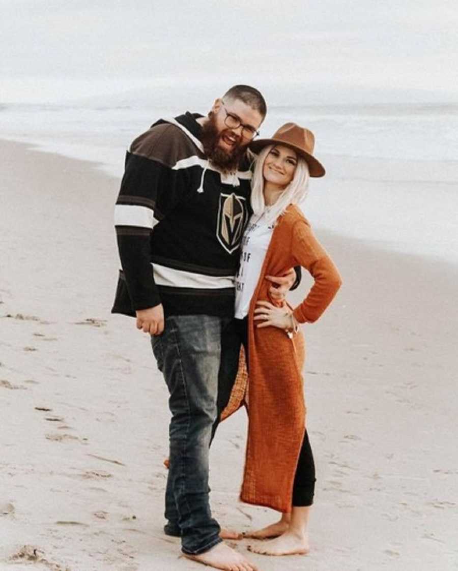 Woman with hodgkin's lymphoma stands smiling on beach in arms of her husband 