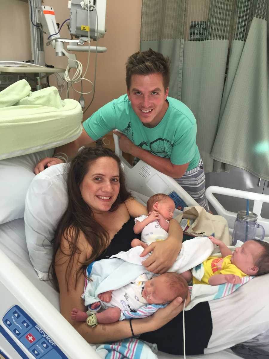 Woman lays smiling in hospital bed as she has triplet newborns in her lap and her husband stands smiling over them