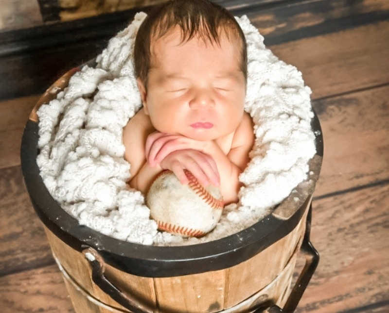 Newborn sits in wooden bucket wrapped in white blanket holding onto baseball
