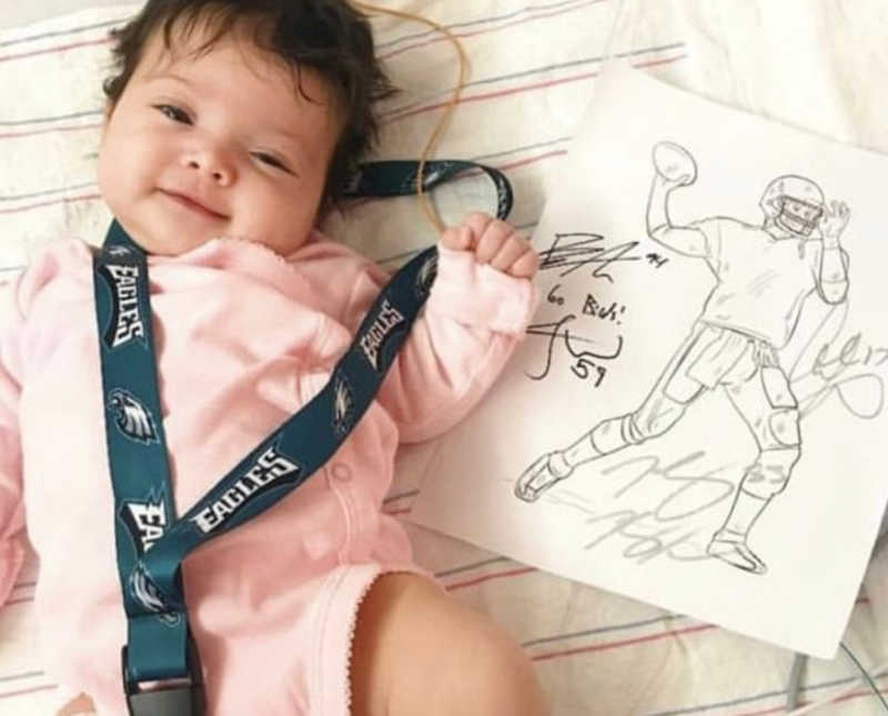 Baby with Spina Bifida lays on back with Philadelphia Eagles lanyard around her beside autographed paper