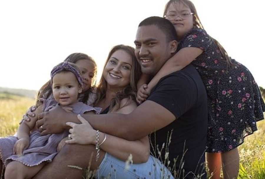 Husband and wife sit in field of grass with their three daughters