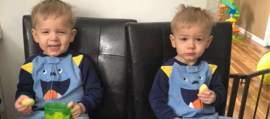 Twin little boys sit in chairs in home in matching onesies eat food