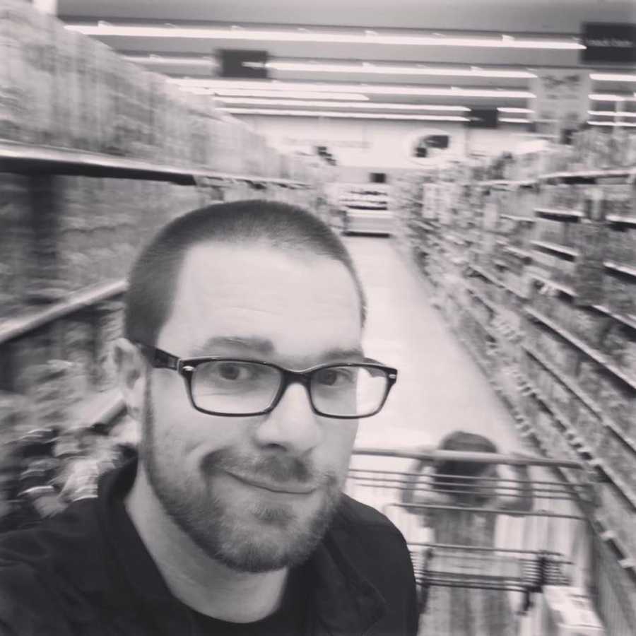 Father takes selfie in grocery store aisle while his little girl pushes shopping cart