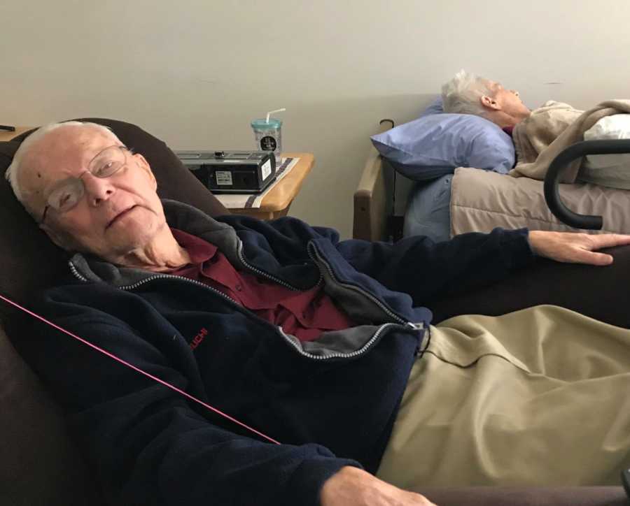 Man lays back in recliner beside his wife with dementia who lays in bed in nursing home