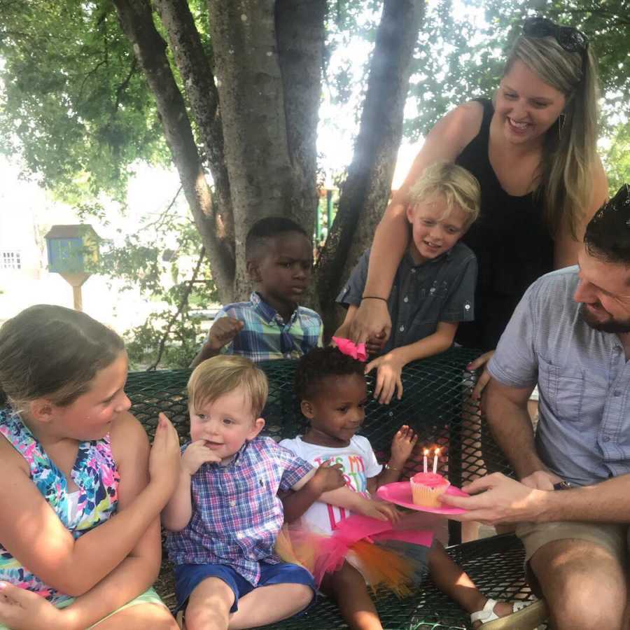 Father sits on bench with three children holding plate with cupcake on it while wife stands behind with their other two children
