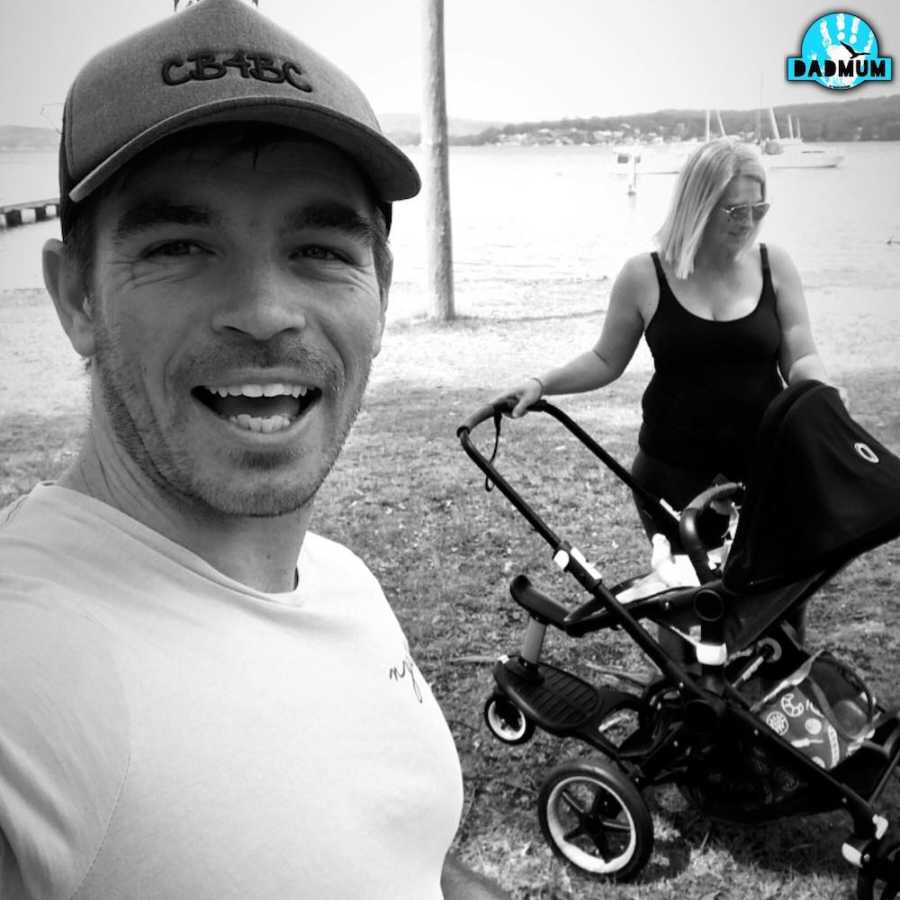 Man smiles in selfie as his wife stands beside stroller in background