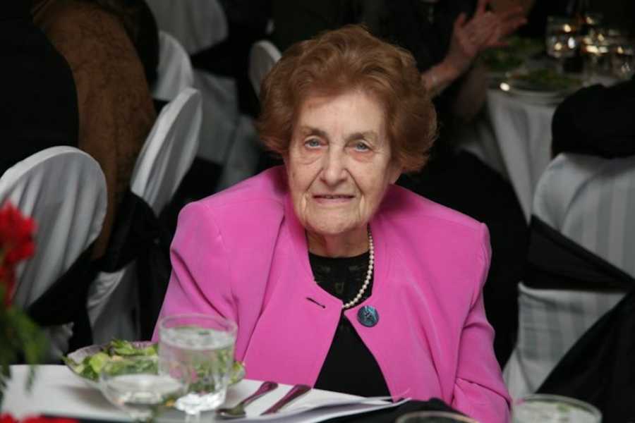 Elderly woman sits smiling at table with pink blazer on 