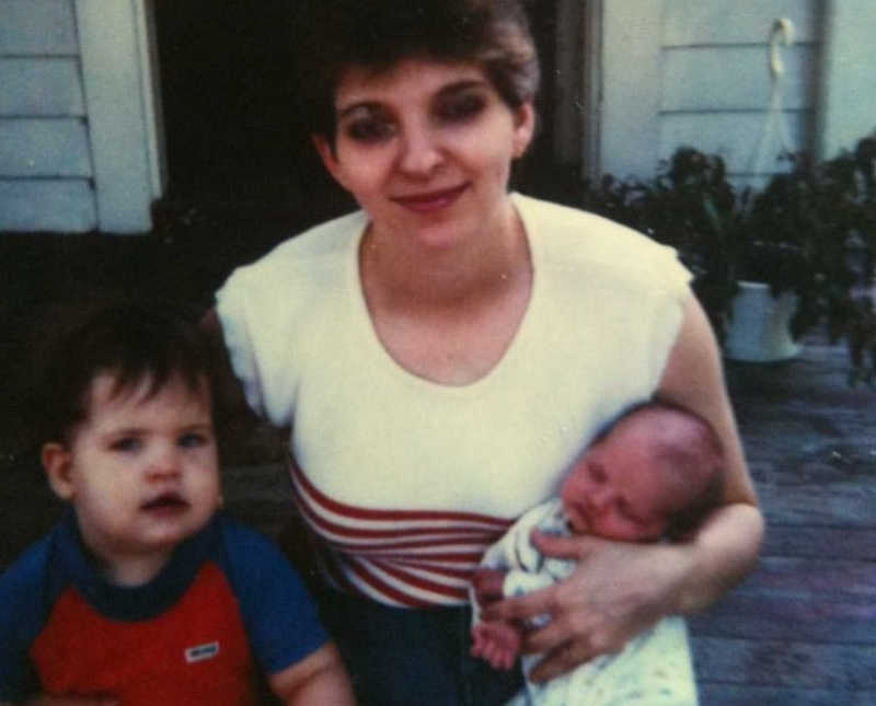 Mother sits outside home with baby in his arm and older son beside her