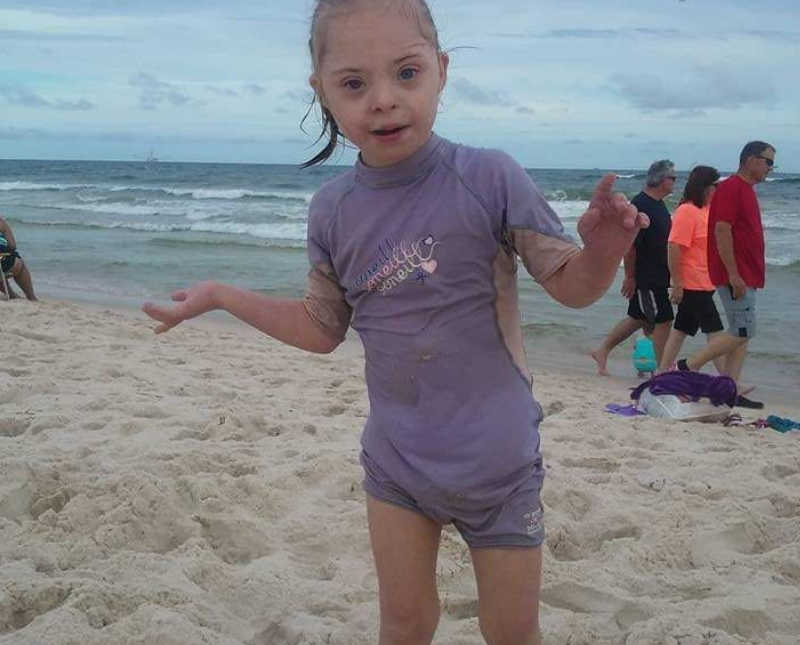 Little girl with down syndrome stands on beach with purple wet suit on