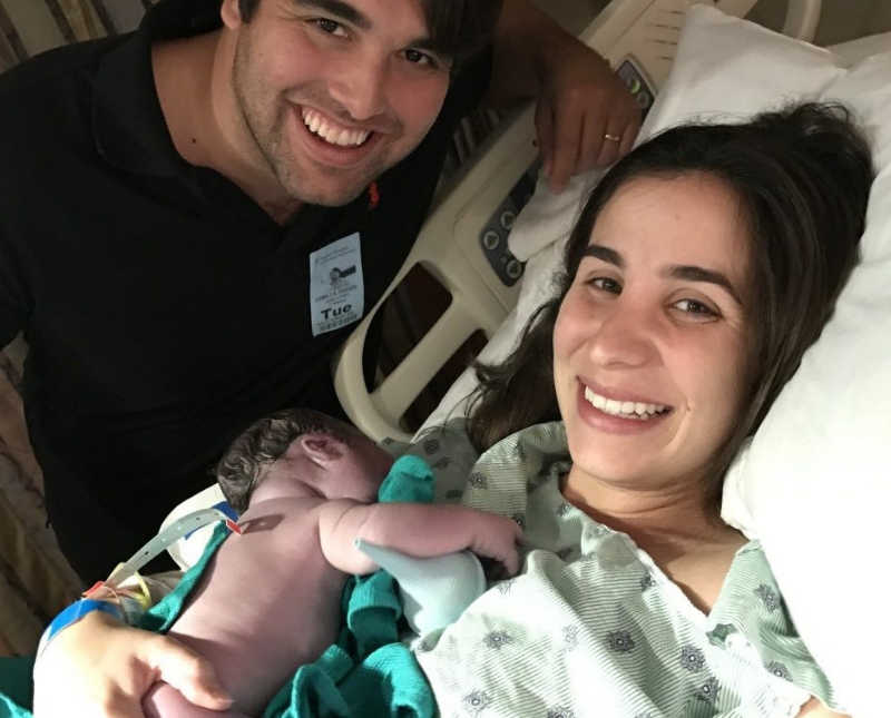 Woman lays in hospital bed holding naked newborn as husband leans over smiling