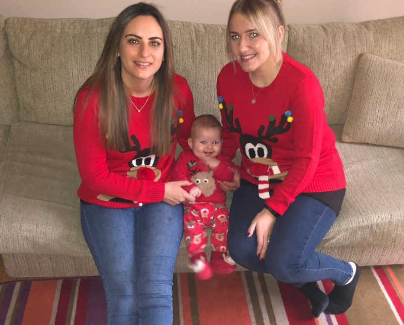 Married woman sit on couch in Christmas sweaters with their baby daughter sitting between them