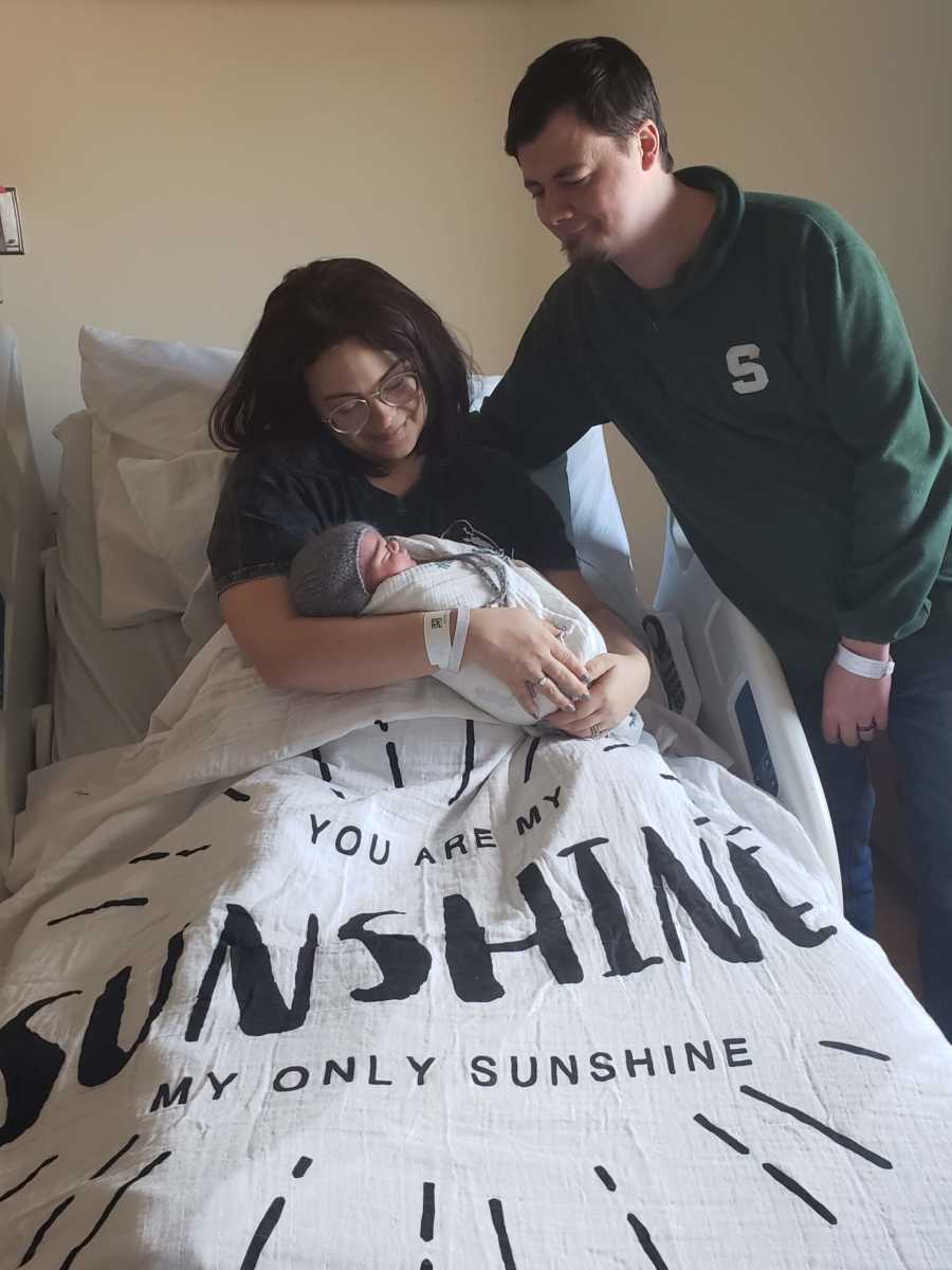Mother smiles in hospital bed holding newborn as husband stands beside her after losing first born