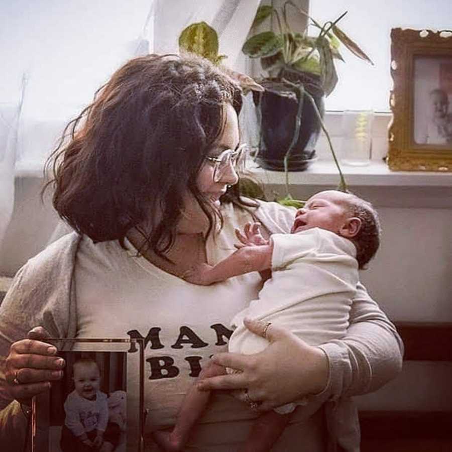 Mother smiles as she holds newborn in one arms and picture frame of deceased new born in other