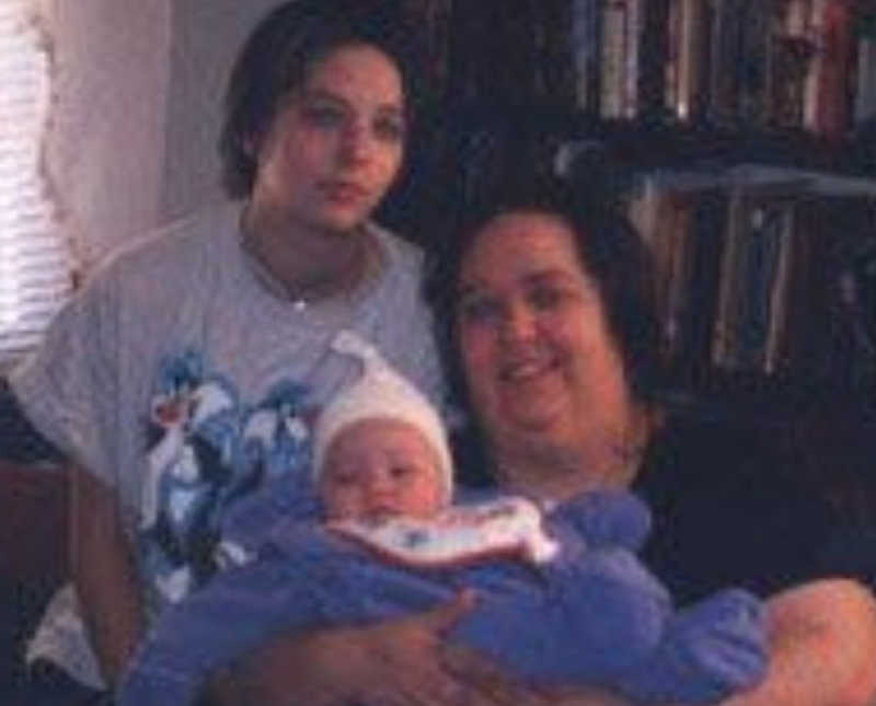 Woman sits in chair holding newborn while teen sits beside her