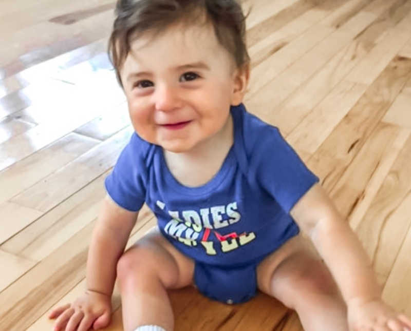 Adopted baby boy sits on floor of home in blue onesie smiling