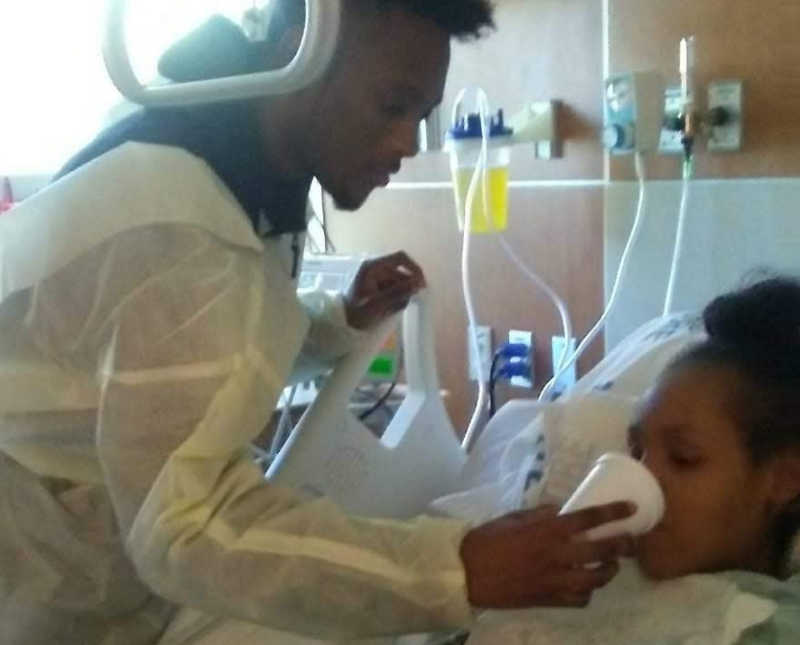 Brother stands tipping cup into sisters mouth who sits in hospital bed