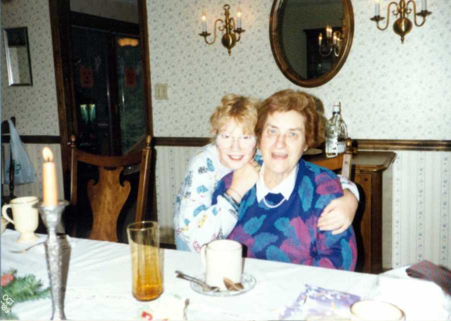 Woman crouches behind grandmother sitting in dining room and hugs her from behind