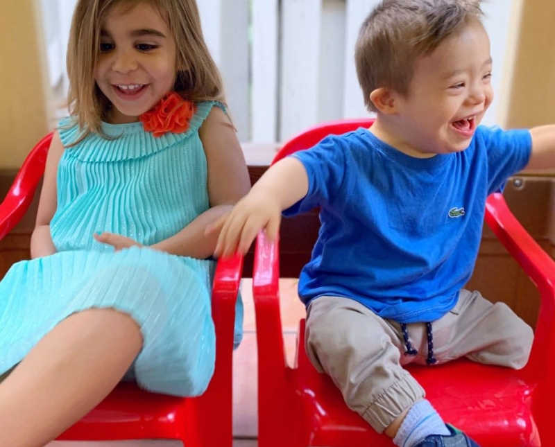 Little boy with down syndrome sits smiling in red plastic chair beside his older sister