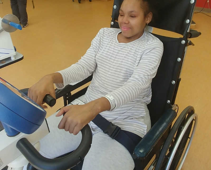 Young woman sits in wheel chair holding on to rehab machine