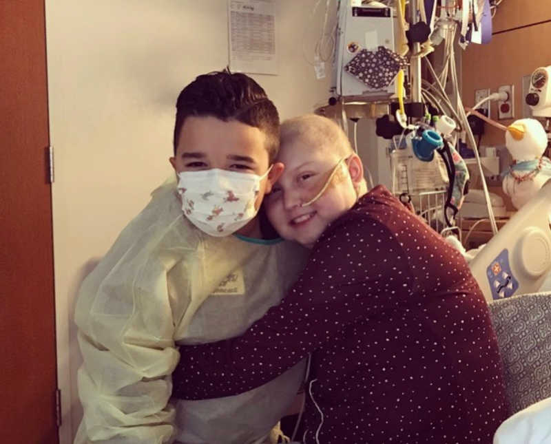 Intubated girl with leukemia sits in hospital bed as she hugs her younger brother with mask on