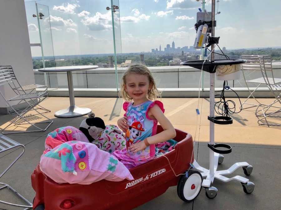 Little girl who is connected to monitor sits on roof top of building in red wagon 