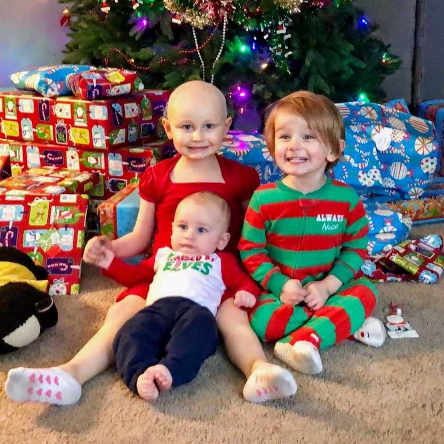 Little girl with leukemia sits on floor with two younger siblings beside presents and Christmas tree