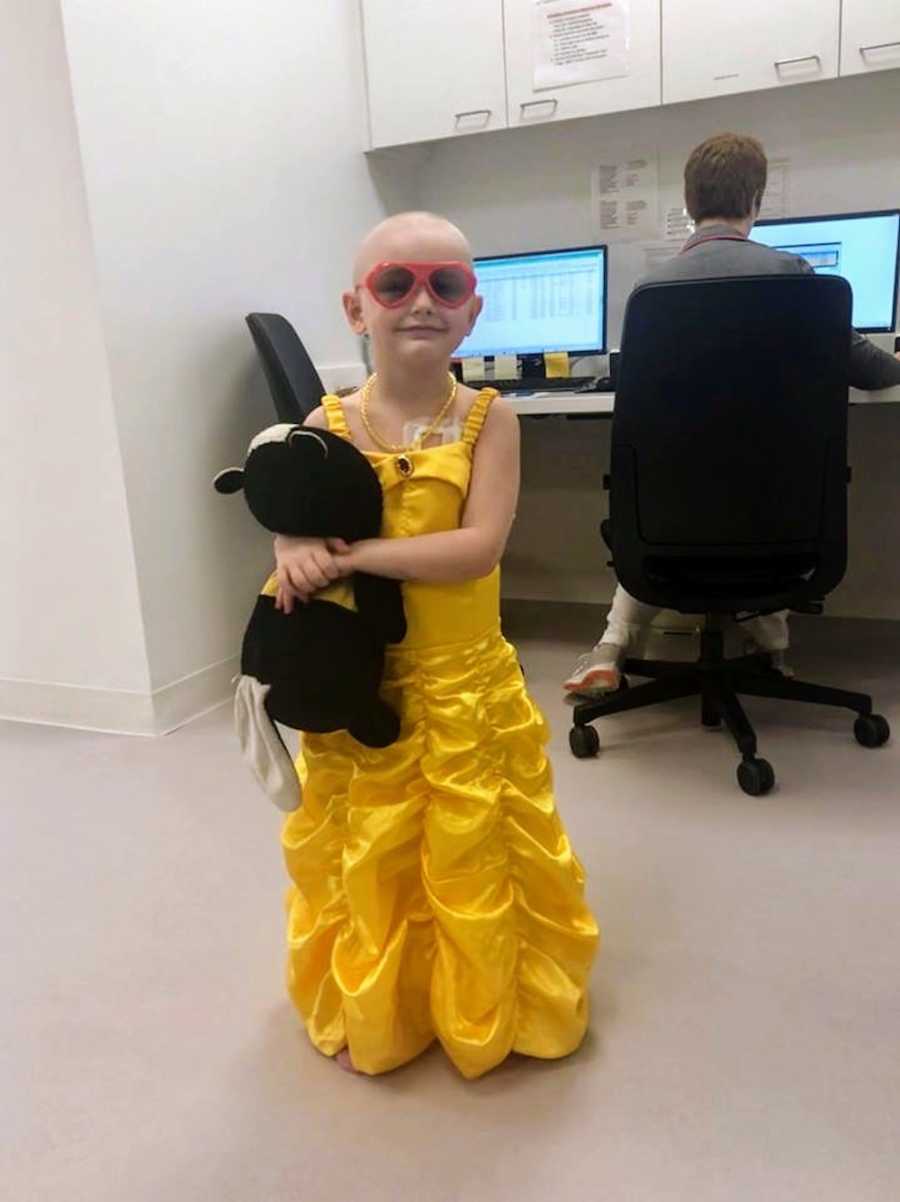 Little girl with Acute Lymphoblastic Leukemia stands in Princess Belle dress holding stuffed animal