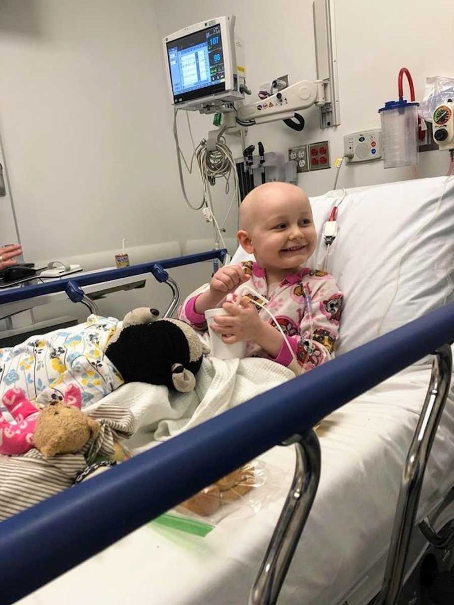 Little girl with Acute Lymphoblastic Leukemia sits smiling in hospital bed with stuffed animals