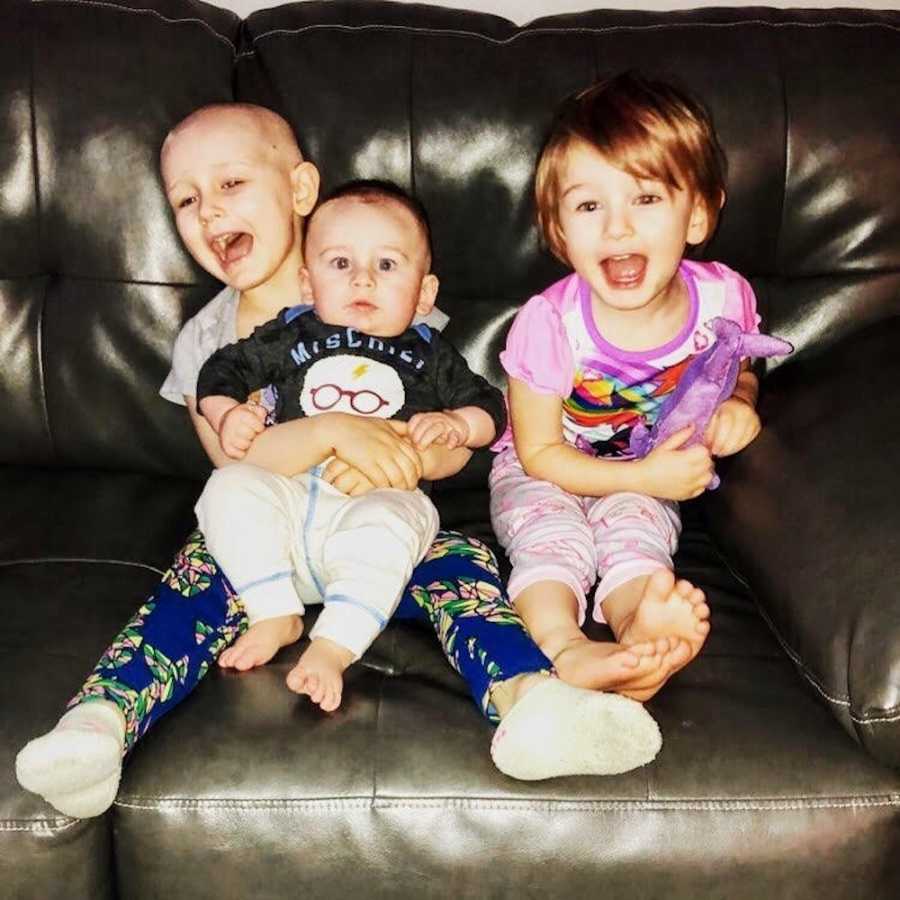 Little girl with Acute Lymphoblastic Leukemia sits on couch with baby brother in her lap and little sister beside her