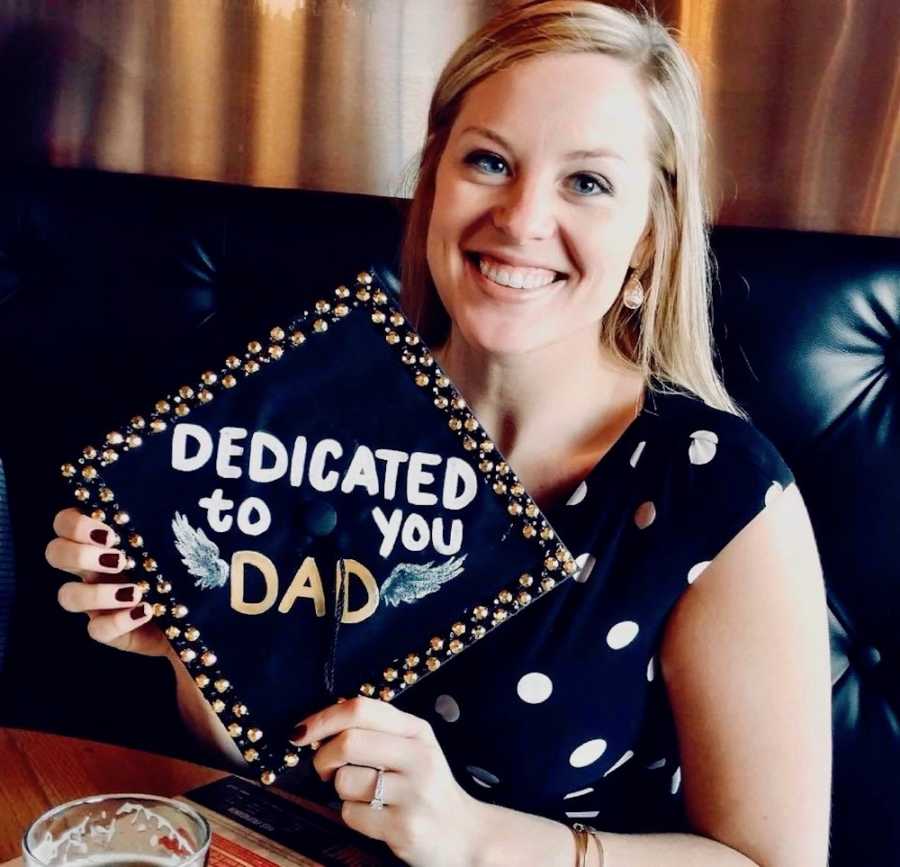 Young woman smiles as she sits on couch holding graduation cap that says, "Dedicated to you Dad"