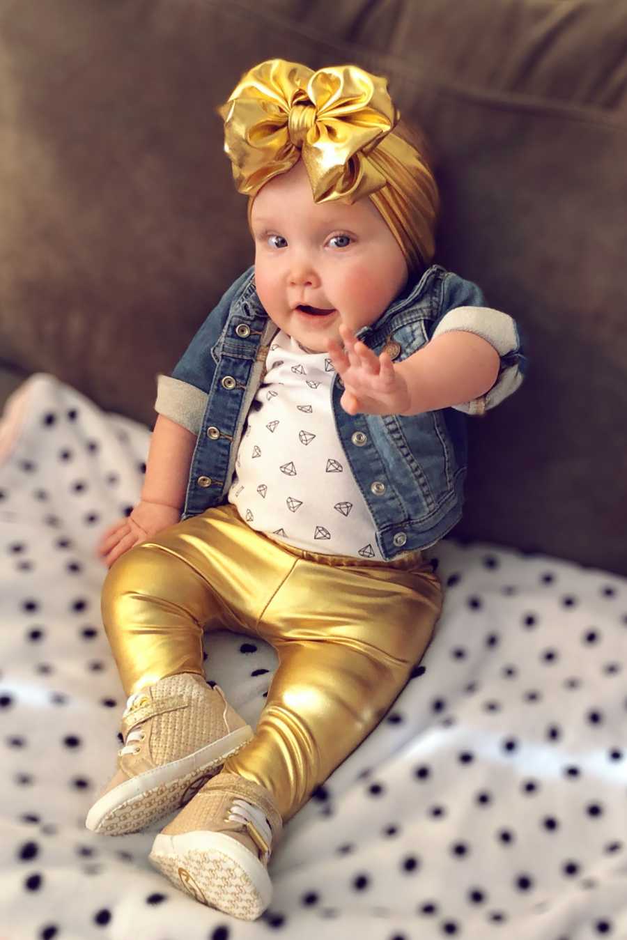 Little girl sits on couch at home with gold legging and matching bow on