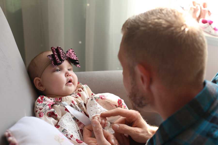 Baby girl sits in chair in home looking at father who crouches in front of her holding his hand
