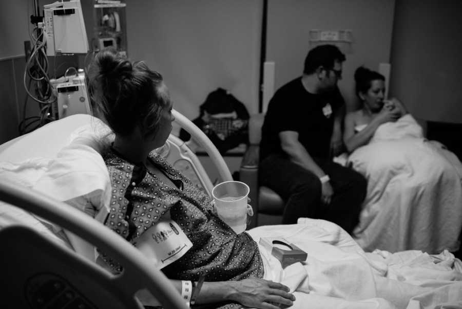 Woman sits up in hospital bed looking at her child that is in arms of adopted mother who sits beside her husband