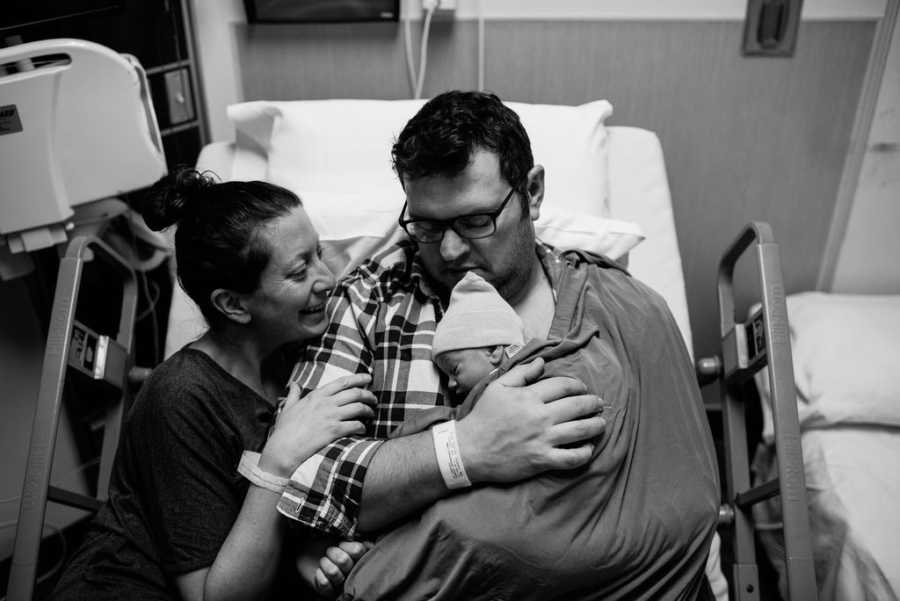 Woman smiles as she lays next to husband in hospital bed who is holding their adopted newborn to his bare chest