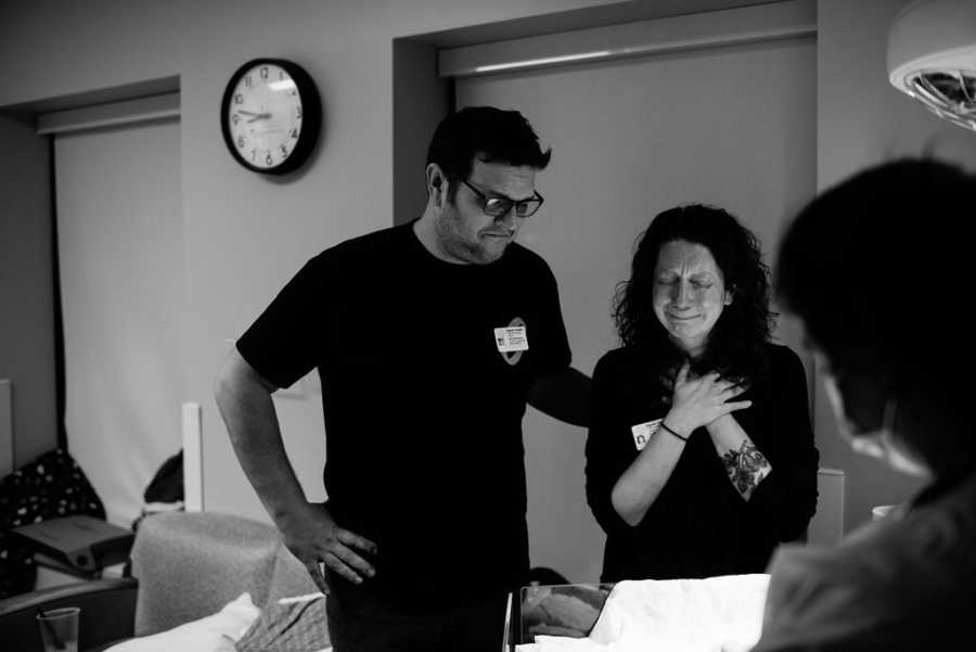 Husband stands with arm on back of wife who stands crying in hospital room