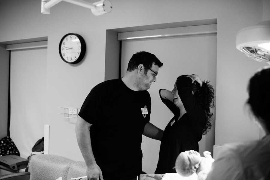 Husband and wife stand in hospital room smiling at each other about birth of their adopted child