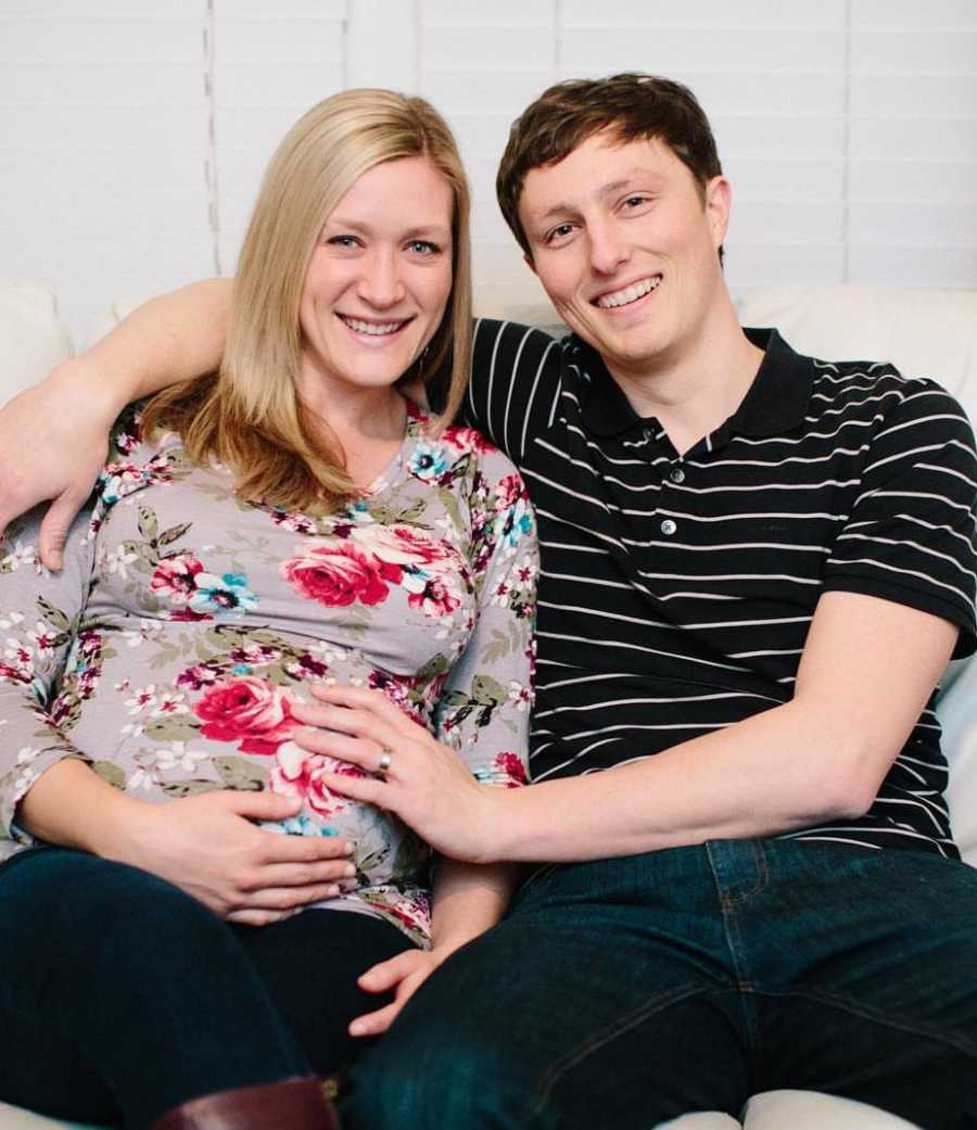Pregnant woman smiles as she sits on couch next to husband who has hand on her stomach