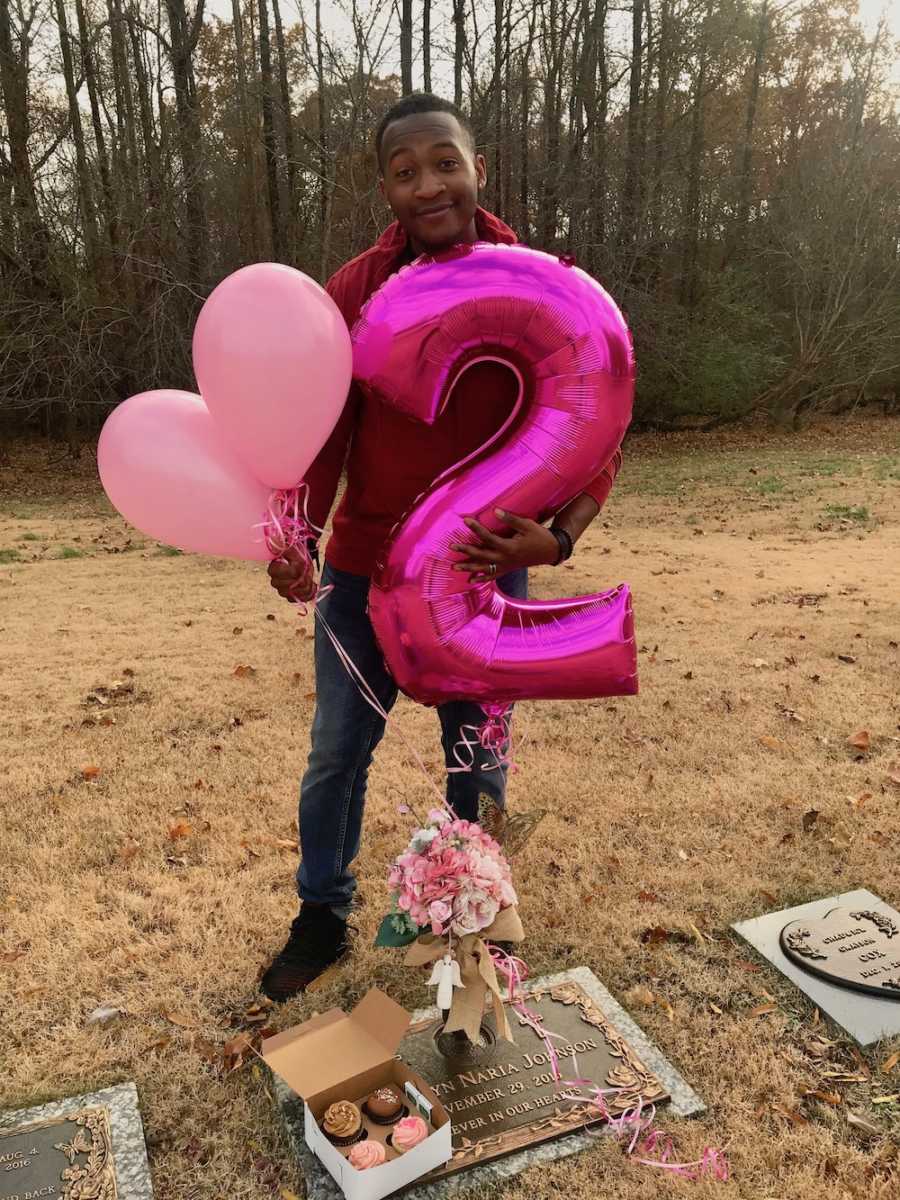 Father stands at hid deceased baby's grave holding pink balloons and pink number two balloon