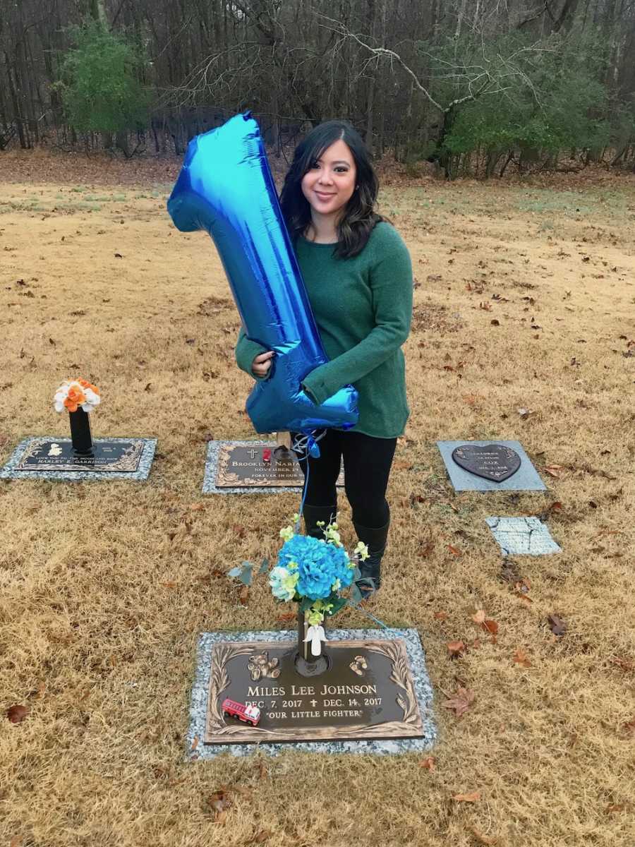 Mother stands at grave of baby who died holding blue number one balloon