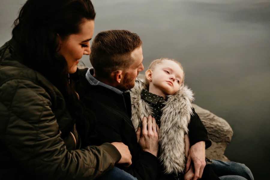 Father sits outside with daughter who has Rett Syndrome in his lap with his wife behind him