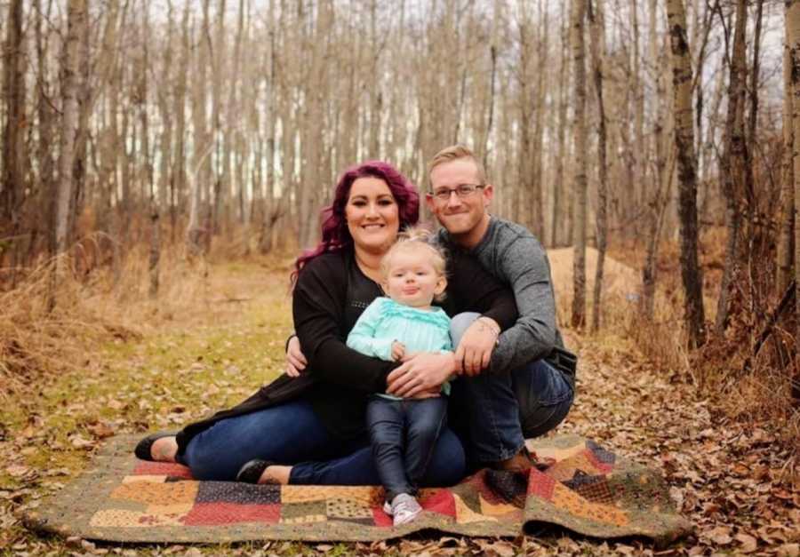 Husband and wife sit on blanket outside in wooden area with their daughter