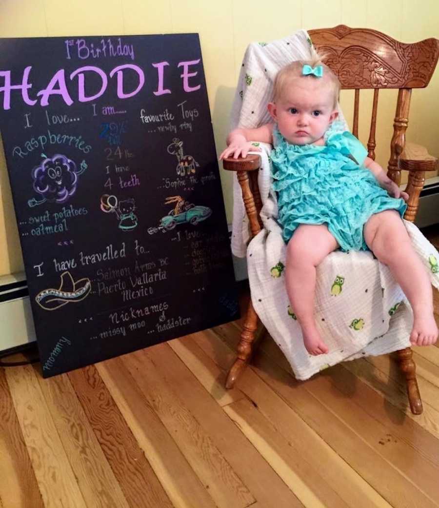 Baby girl sits in wooden chair in home beside sign for her first birthday
