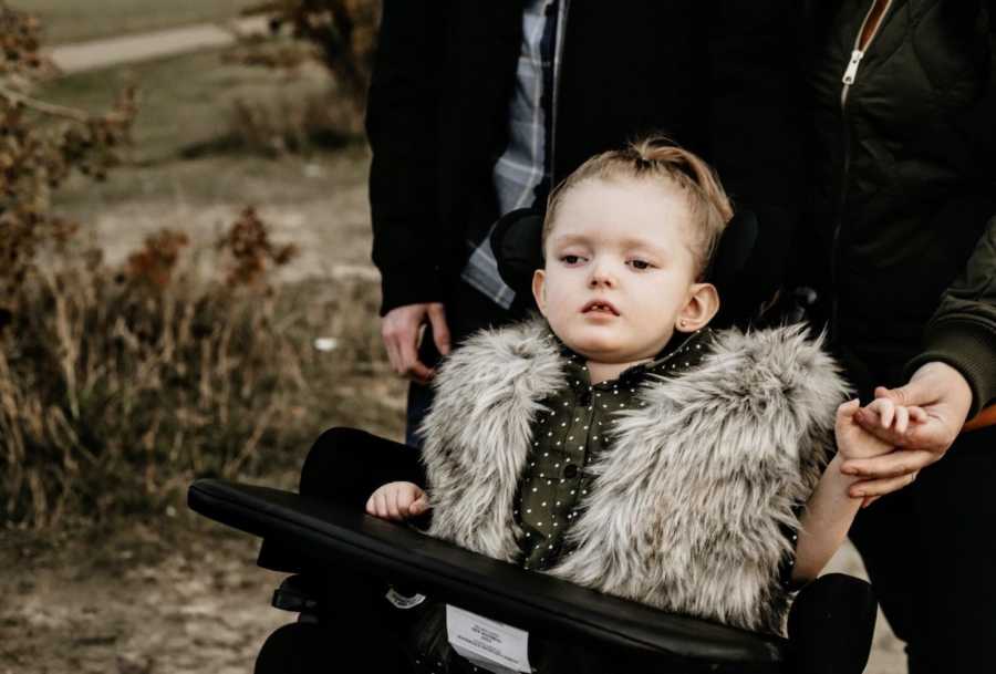 Little girl with Rett Syndrome sits in wheel chair holding mothers hand