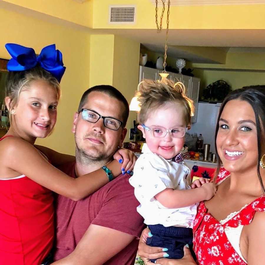 Husband and wife stand smiling in home while husband holds daughter and wife holds son with down syndrome
