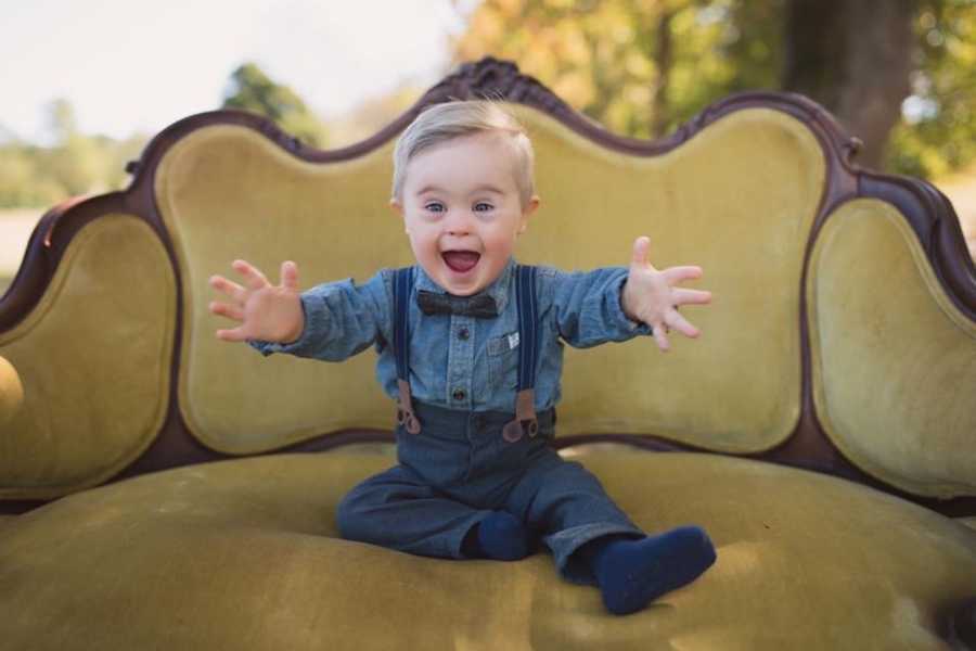 Little boy with down syndrome sits on green velvet couch with mouth open and arms out to his side